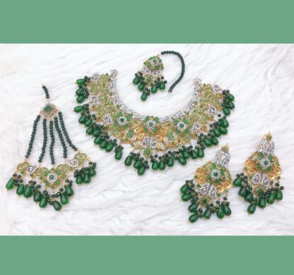 A luxurious gold-plated Kundan necklace set with green emerald beads, intricate polki stones, matching earrings, a maang tikka, and a passa, displayed on a soft fabric background.