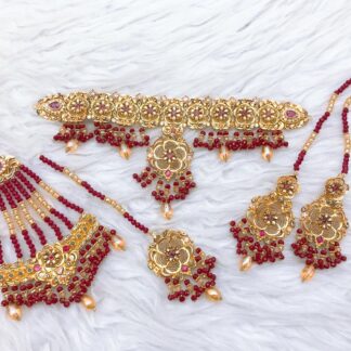 A handcrafted gold-plated Kundan choker set featuring intricate floral motifs, deep red ruby bead embellishments, matching earrings, a maang tikka, and a haath phool, perfect for brides and special occasions.