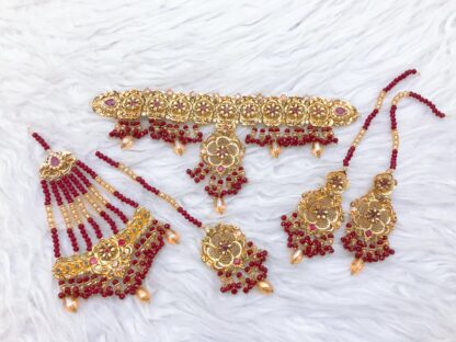 A handcrafted gold-plated Kundan choker set featuring intricate floral motifs, deep red ruby bead embellishments, matching earrings, a maang tikka, and a haath phool, perfect for brides and special occasions.