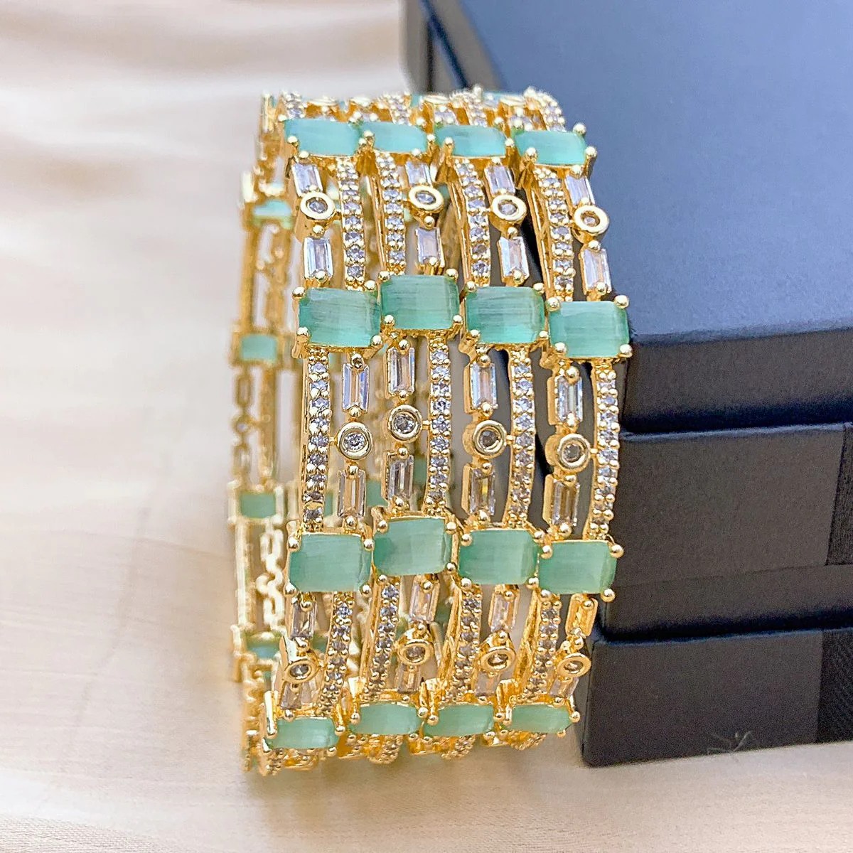 Mint Green AD Bangles (Set of 4) Overlapping gold-plated bangles with alternating mint green and clear stones, arranged on a velvet-textured jewelry box for catalog photography.