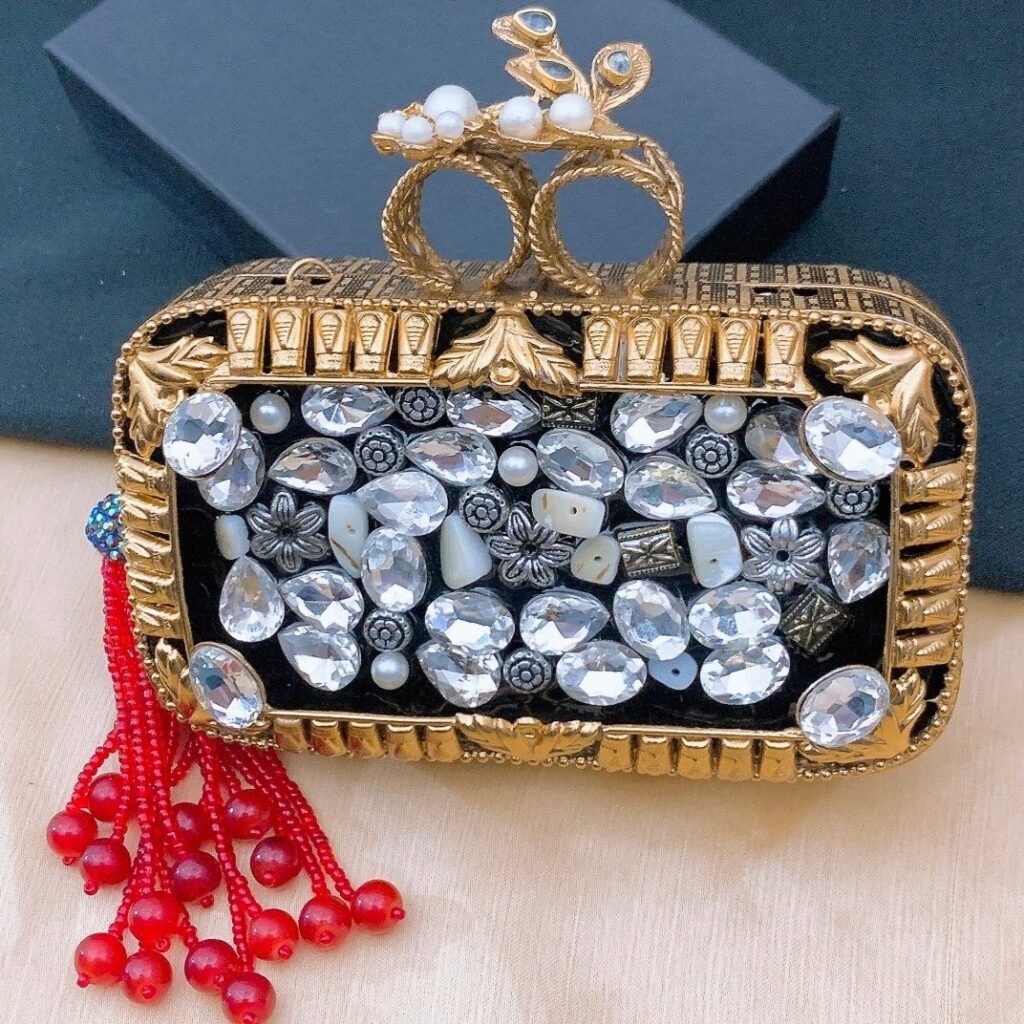 An elegant, hard-case evening clutch (minaudière) covered in white faux pearls and clear silver crystals set on a dark/black background. The bag features a decorative gold knuckle ring closure with a bee/floral motif and a striking red beaded tassel for a formal event or bridal look.