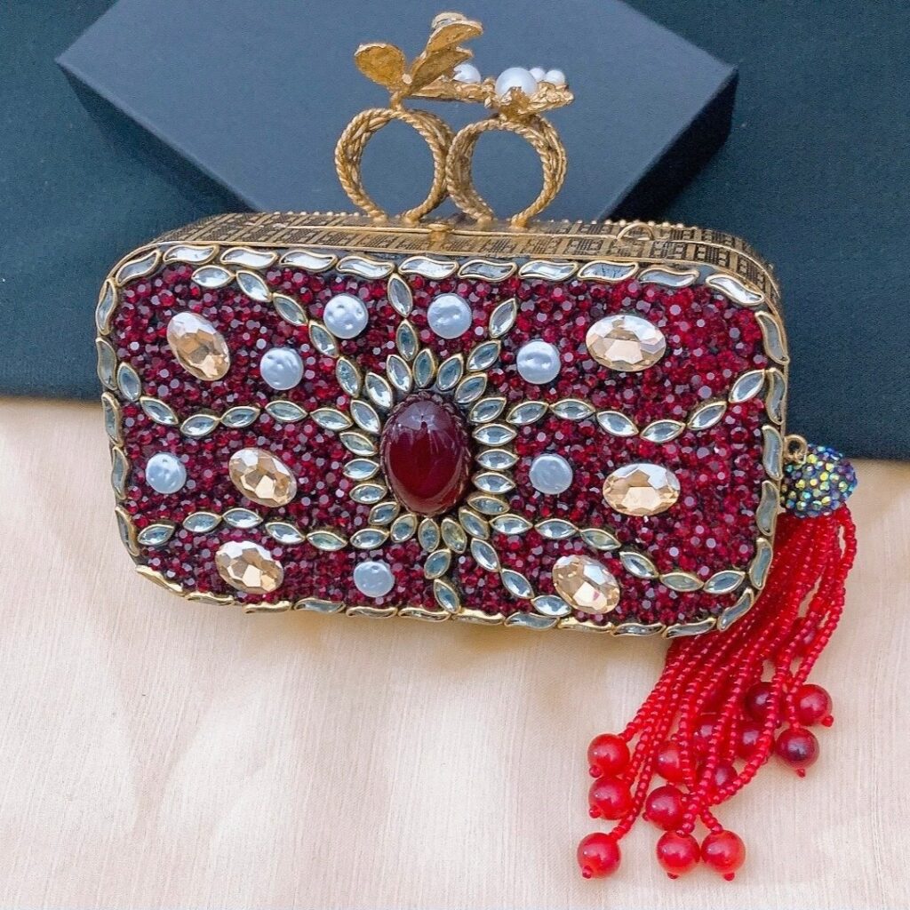 A stunning, regal burgundy evening clutch (minaudière) covered in deep red, clear, and champagne crystals and pearls. It has a decorative gold knuckle ring closure with a bee/floral motif and a long, vibrant red beaded tassel attached to the side.