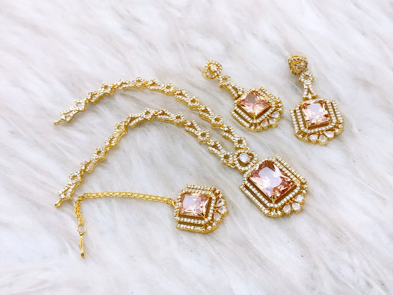 Peach Champagne Radiant-Cut Luxury Jewelry Set 4-piece peach champagne jewelry set featuring a gold-plated necklace, earrings, and maang tikka on a white faux fur background.