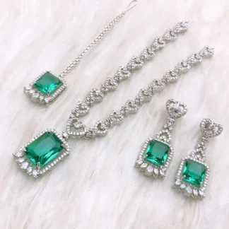 4-piece emerald green and silver jewelry set featuring a necklace, drop earrings, and maang tikka on a white faux fur background.