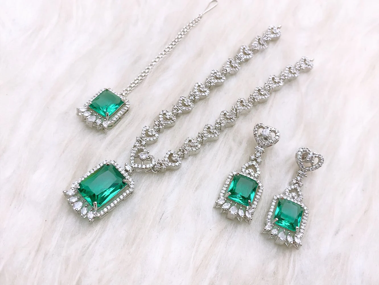 Emerald Green Radiant-Cut Luxury Jewelry Set 4-piece emerald green and silver jewelry set featuring a necklace, drop earrings, and maang tikka on a white faux fur background.