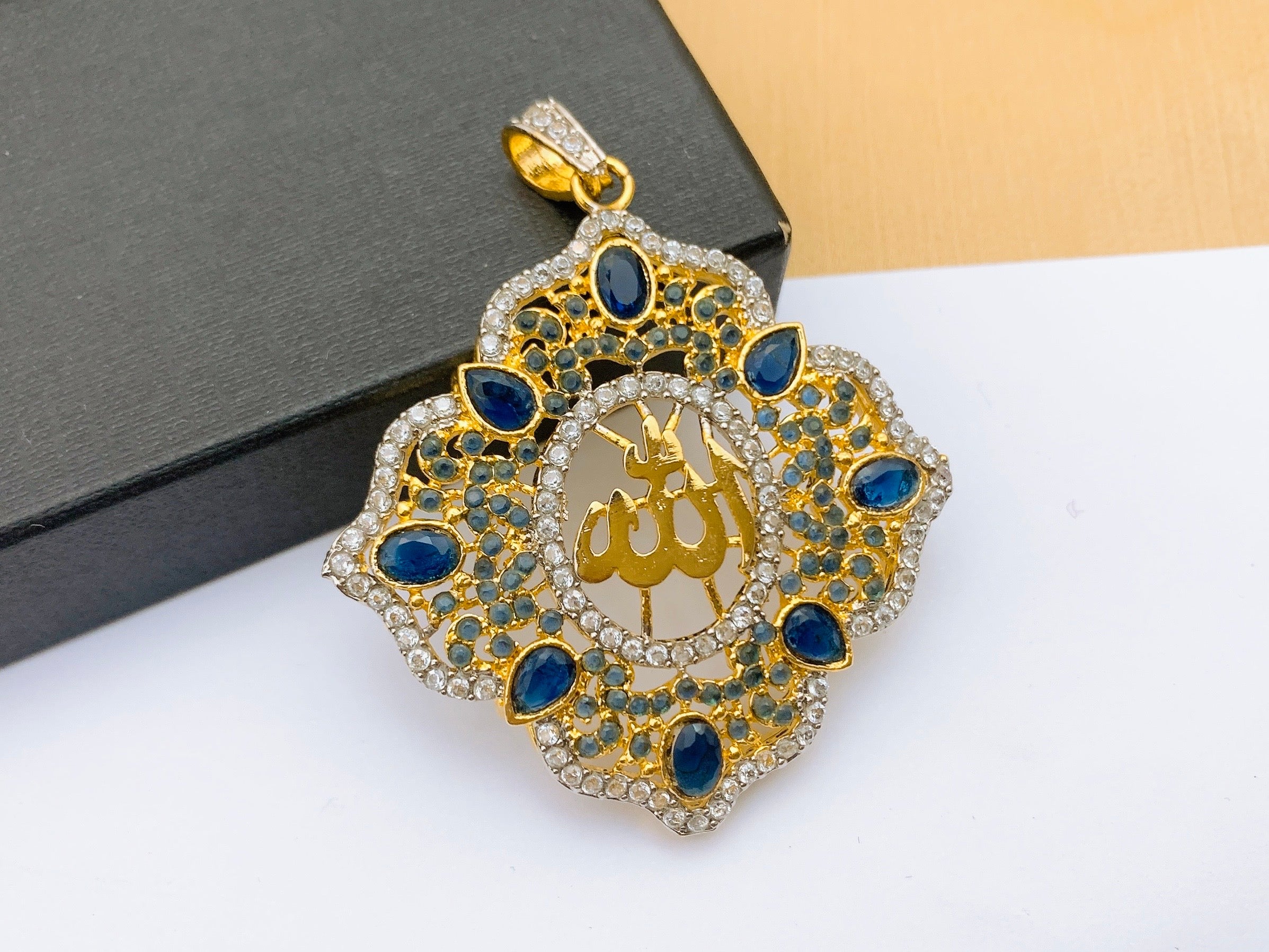 Majestic Sapphire Blue Allah Calligraphy Pendant with Diamond Halo A majestic sapphire blue Allah calligraphy pendant with gold script, featuring an ornate scalloped border set with blue crystals and brilliant white diamonds.